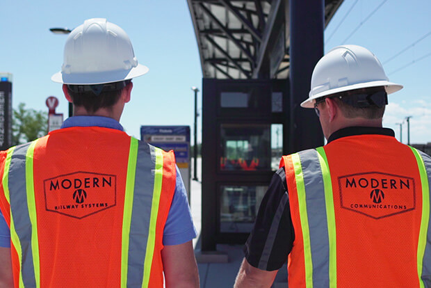 Two workers in vests displaying the Modern Railways Logo.